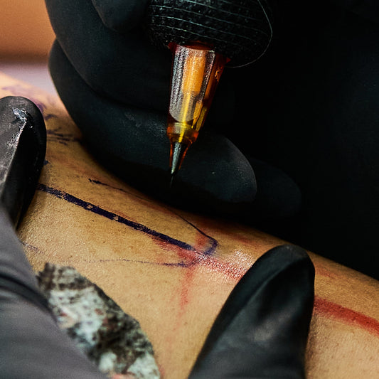 Macro view of a fine liner tattoo needle precisely tracing a purple stencil on skin.