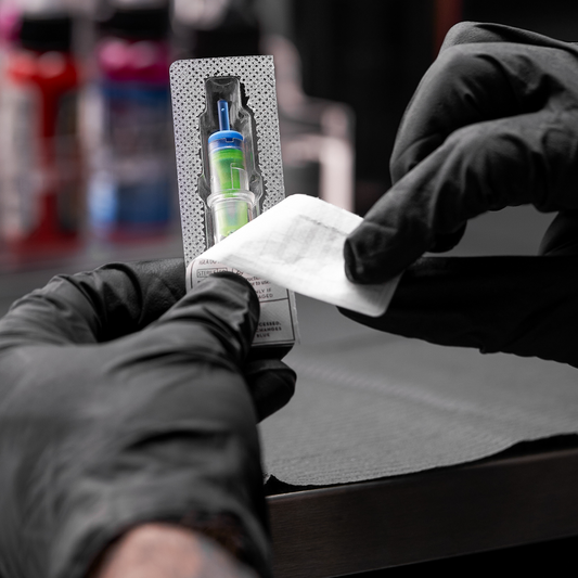 A tattoo artist wearing black gloves peeling open the sterile blister packaging of a new tattoo needle cartridge.