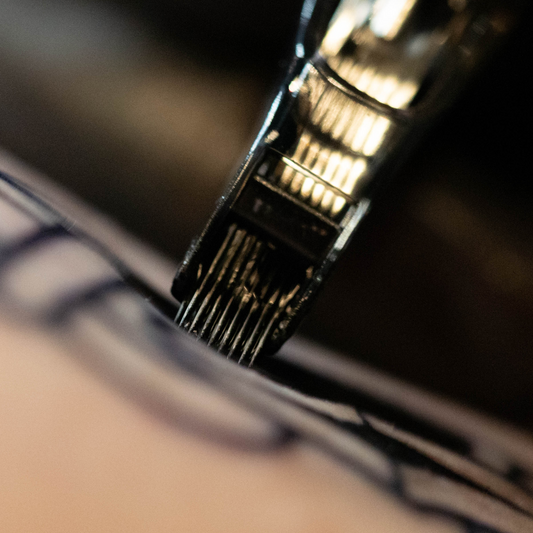 A dramatic, high-contrast close-up focusing on the exposed metallic tips of a flat magnum tattoo needle grouping just above a dark stenciled line.