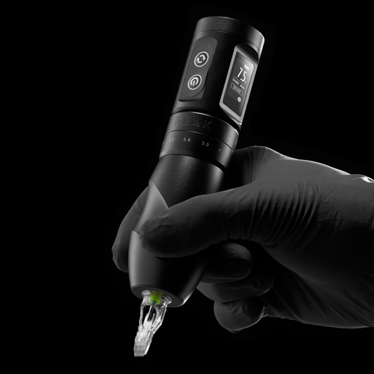 A gloved hand holding a black Peak wireless tattoo machine with a digital side display.