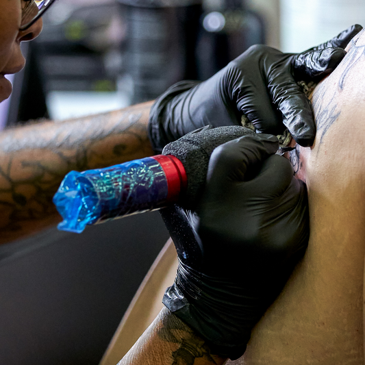 A close-up of a tattoo artist wearing black gloves while using a rotary machine to tattoo a client's skin.